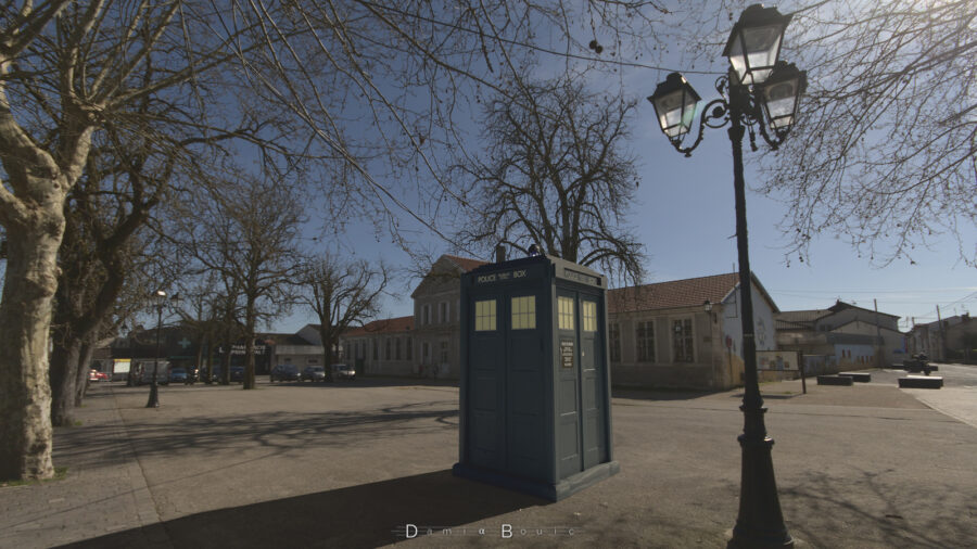 Le TARDIS sur une place type village français, avec des marroniers dépourvus de leur feuillages, une école primaire en arrière plan, un lampadaire à droite