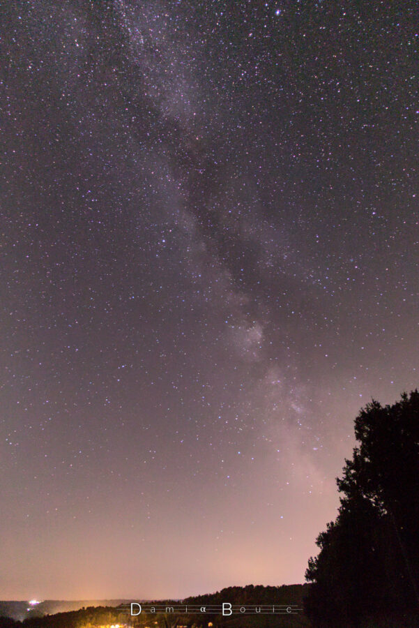 Ciel nocturne, avec la Voie Lactée qui barre le ciel depuis l'horizon vers le zénith. Quelques lumières d'un petit village sont visibles en bas, et un halo lumineux en partie caché par un arbre tout en bas à droite