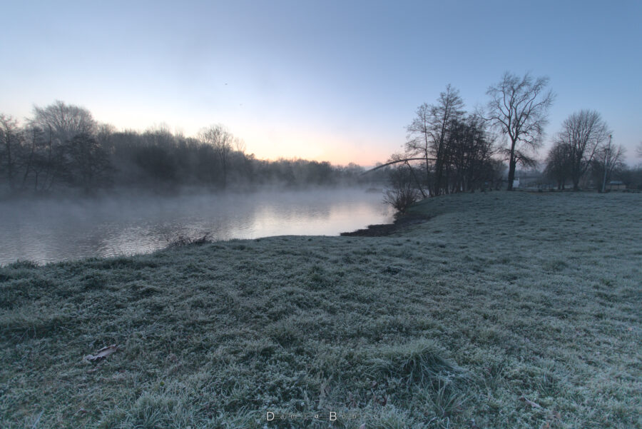 Ambiance froide et hivernale, le givre recouvre la pelouse. A l'arrière, la rivière est fumante, et contraste vivement avec une forêt d'arbres encore décharnées